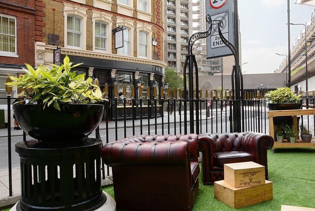 Hotel Montcalm Brewery Townhouse Londyn Zewnętrze zdjęcie The photo shows a street scene with a cozy outdoor seating area. There are two plush, dark leather couches arranged on green grass, creating a welcoming atmosphere. Surrounding the seating are large planters with vibrant plants. In the background, th