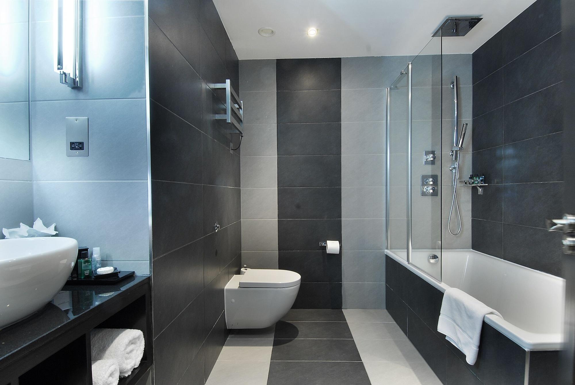 Hotel Montcalm Brewery Townhouse Londyn Zewnętrze zdjęcie The photo shows a modern bathroom featuring a sleek design. The walls are tiled in a combination of dark gray and light gray, creating a contemporary look. On the left, there is a countertop with a basin and some toiletries placed on it. The middle s