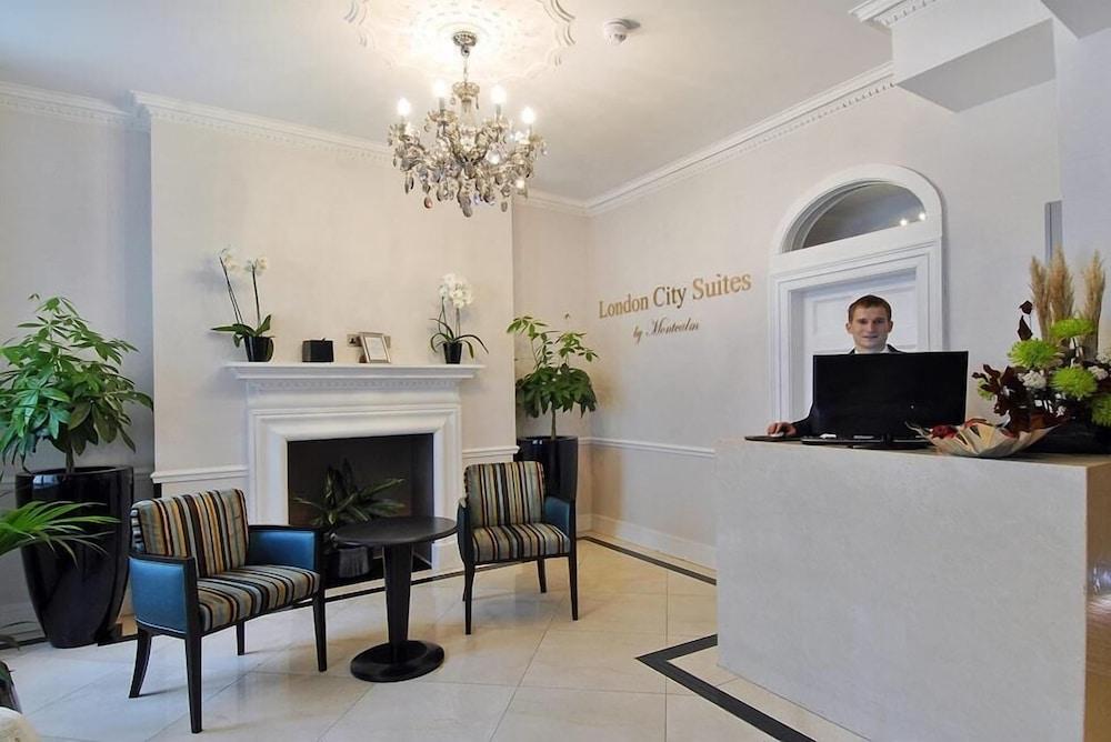 Hotel Montcalm Brewery Townhouse Londyn Zewnętrze zdjęcie The photo shows the interior of a reception area in a hotel called "London City Suites." The space features a light-colored wall with the hotel's name displayed. There is a stylish chandelier hanging from the ceiling, providing warm lighting. 

In th