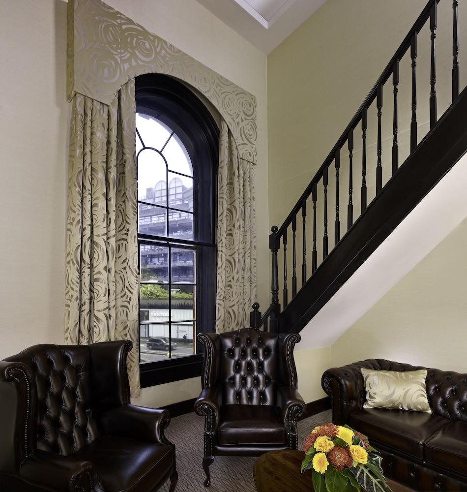 Hotel Montcalm Brewery Townhouse Londyn Zewnętrze zdjęcie The photo shows a cozy interior space featuring two elegant leather armchairs placed near a large, ornate window with arched top and decorative curtains. The wall is painted in a muted color, enhancing the warmth of the room. A staircase with black s