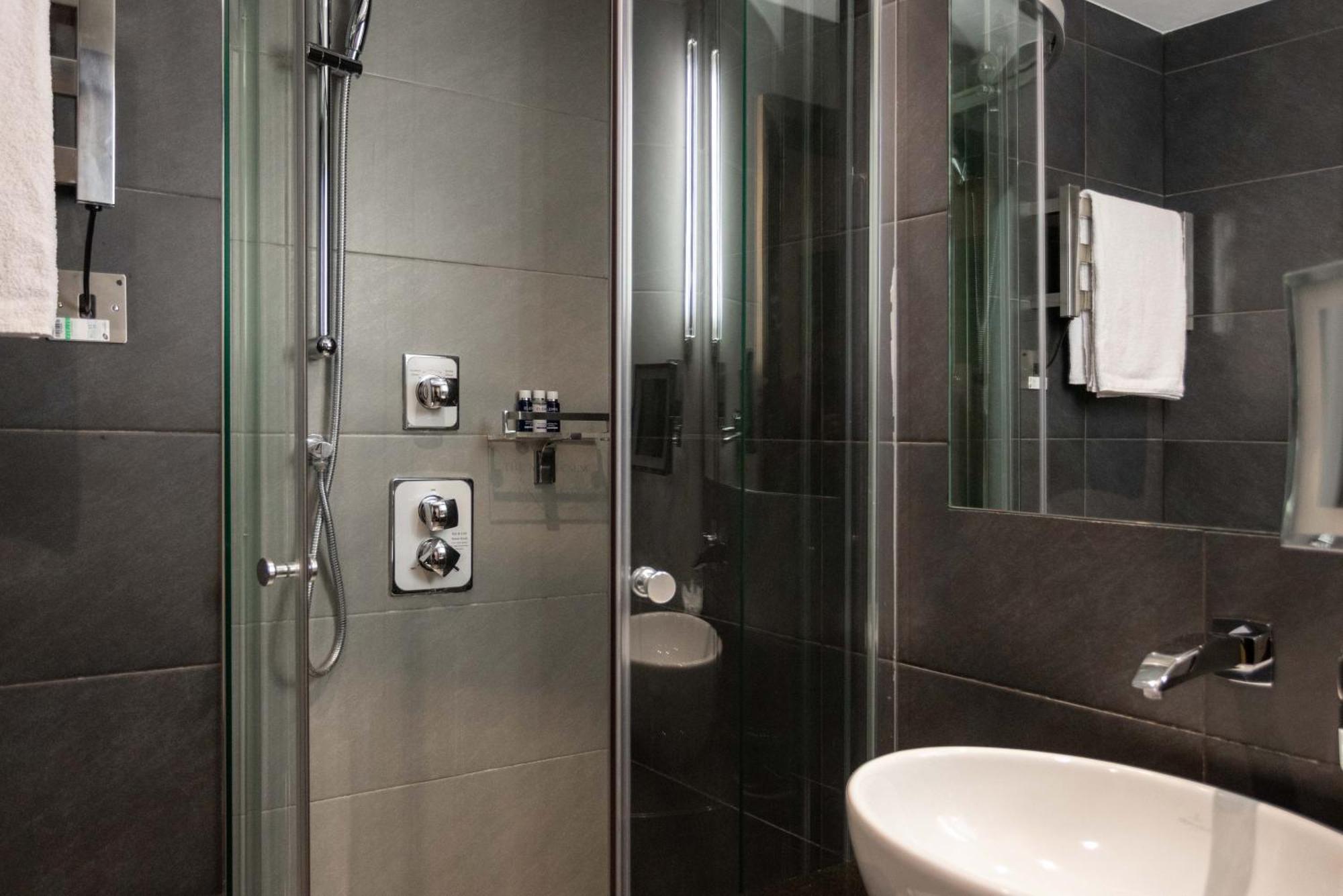 Hotel Montcalm Brewery Townhouse Londyn Zewnętrze zdjęcie The photo shows a modern bathroom with a glass shower enclosure. The shower features a handheld showerhead and various controls mounted on the wall. The walls are tiled in dark, sleek tones, contributing to a contemporary aesthetic. There is a round 