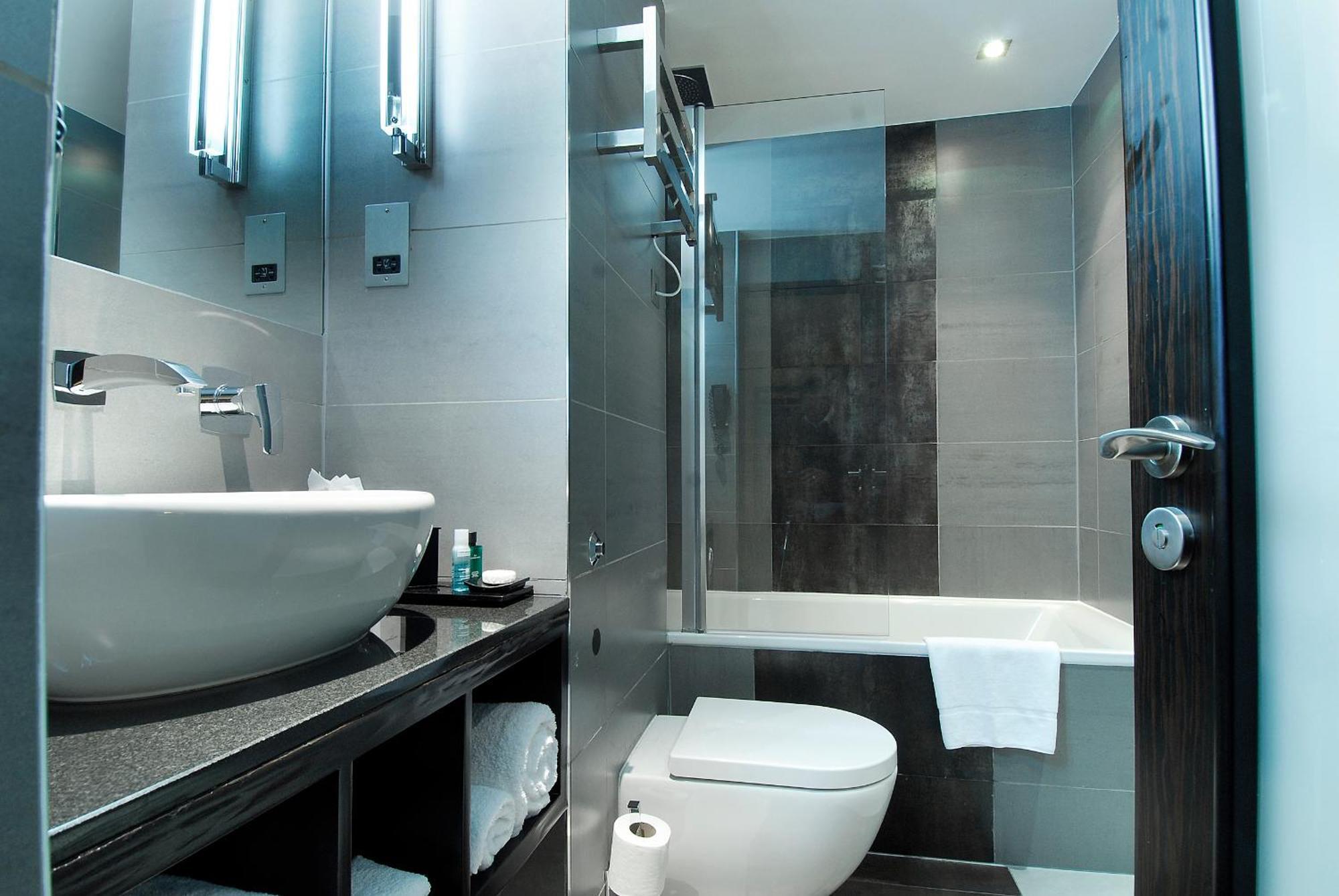 Hotel Montcalm Brewery Townhouse Londyn Zewnętrze zdjęcie The photo shows a modern bathroom with sleek design elements. It features a curved white sink on a dark countertop, with a mirror above that is illuminated by stylish lighting fixtures. Next to the sink, there's a toilet, and on the opposite side, a 