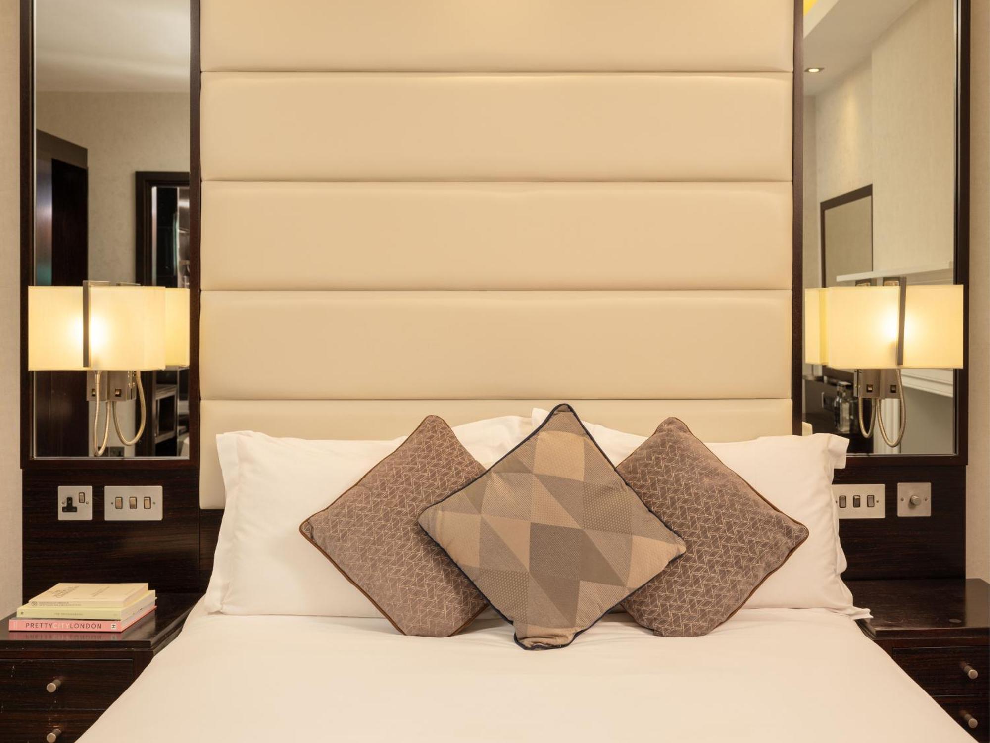 Hotel Montcalm Brewery Townhouse Londyn Zewnętrze zdjęcie The image shows a cozy bedroom setting. The bed features a headboard upholstered in a light beige fabric. It is adorned with three decorative pillows in varying patterns and colors, placed on white bedding. On either side of the bed, there are modern