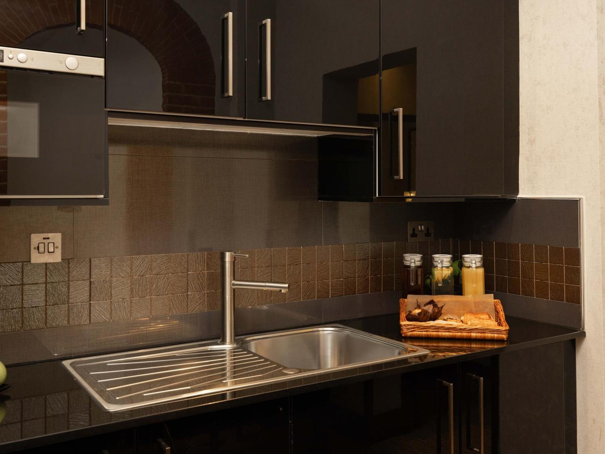 Hotel Montcalm Brewery Townhouse Londyn Zewnętrze zdjęcie The photo shows a modern kitchen with a sleek design. It features dark cabinetry with a glossy finish and stainless steel handles. There is a stainless steel sink with a simple faucet, and a countertop made of black material. Above the sink, there ar