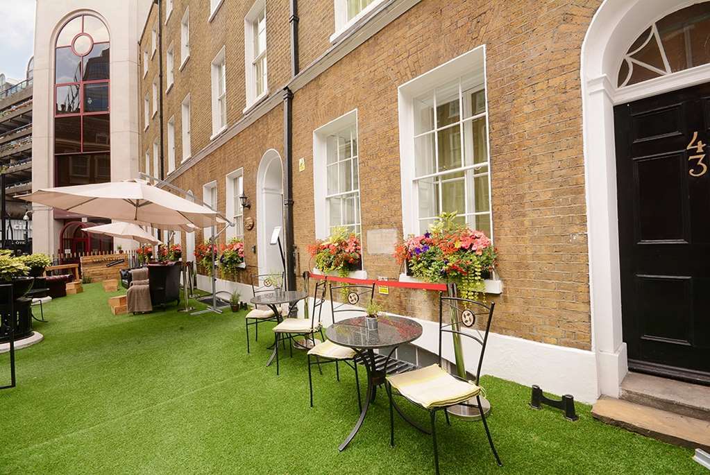 Hotel Montcalm Brewery Townhouse Londyn Zewnętrze zdjęcie The photo depicts a charming outdoor area in front of a building with a warm, textured brick facade. There is a neatly maintained lawn, likely made of artificial grass, and several small round tables with chairs are arranged outside. Flower pots with