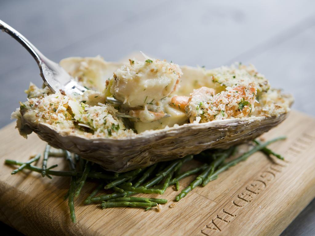 Hotel Montcalm Brewery Townhouse Londyn Zewnętrze zdjęcie The photo shows a large oyster shell filled with a mixture that appears to include seafood, herbs, and breadcrumbs. The dish is presented on a wooden board with some greenery, possibly a type of seaweed or garnishing. A fork is placed beside the oyst