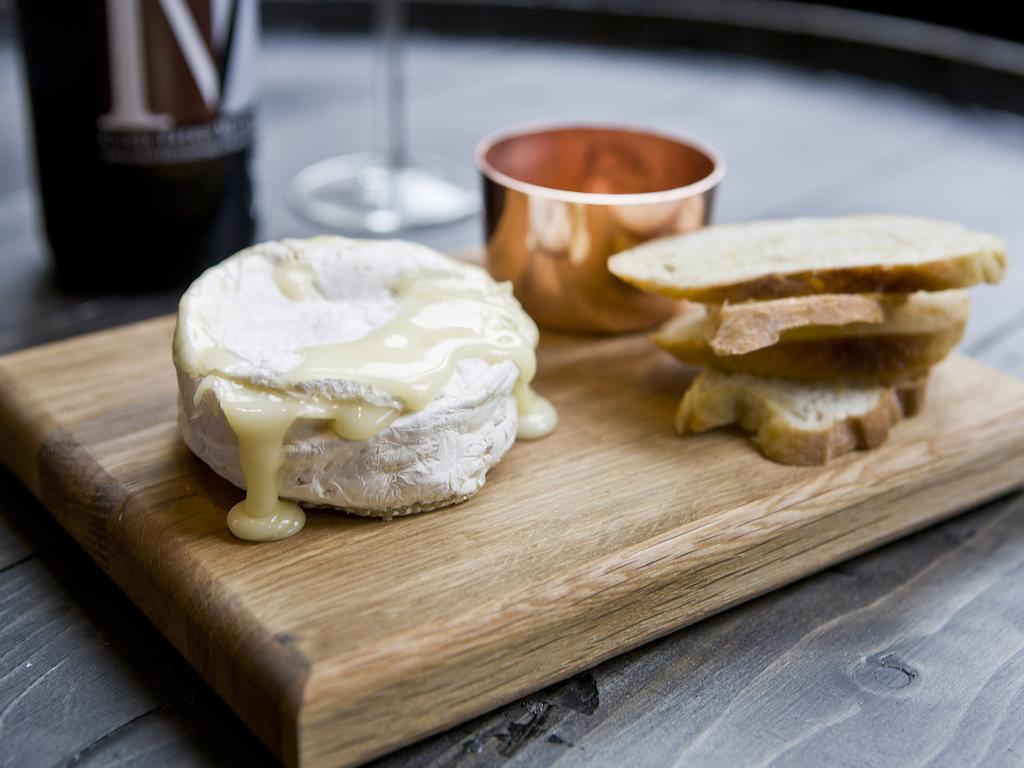 Hotel Montcalm Brewery Townhouse Londyn Zewnętrze zdjęcie The photo features a wooden serving board with several items. There is a round cheese, likely Brie or a similar soft cheese, that appears to be melting and dripping slightly. Next to the cheese, there is a small copper cup that might contain a sauce 