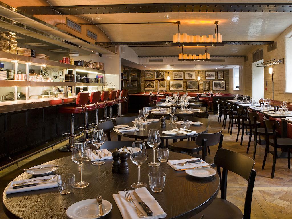 Hotel Montcalm Brewery Townhouse Londyn Zewnętrze zdjęcie The photo shows the interior of a stylish restaurant. It features several round tables set with white plates, glasses, and silverware, all neatly arranged for dining. The decor includes a modern design with wooden floors and a mix of lighting fixture