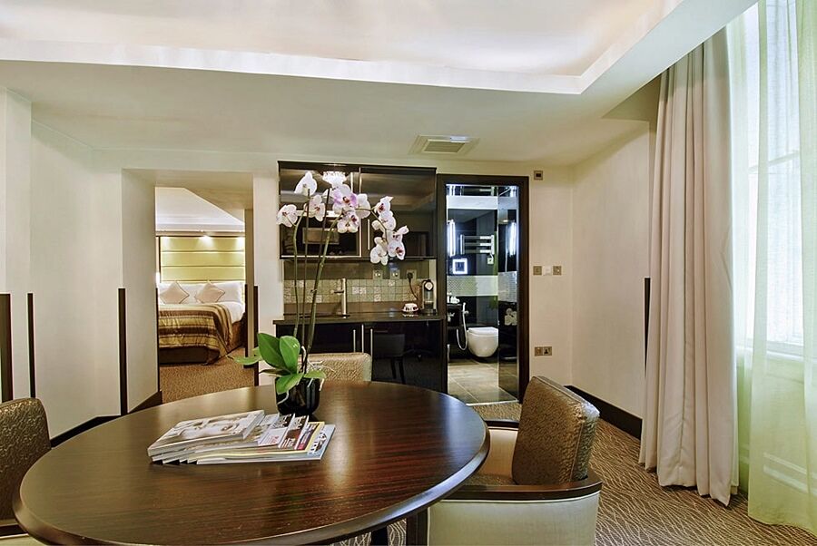 Hotel Montcalm Brewery Townhouse Londyn Zewnętrze zdjęcie The photo shows a stylish hotel room or suite. In the foreground, there is a round dining table with a few magazines on it. Surrounding the table are upholstered chairs. To the right, there are large windows with light-colored curtains, allowing natu
