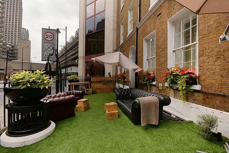 Hotel Montcalm Brewery Townhouse Londyn Zewnętrze zdjęcie The photo depicts a charming outdoor space, likely a patio or garden area. It features two comfortable-looking leather sofas, one of which is a deep burgundy color, placed on a green artificial lawn. There are wooden crates that could serve as side t